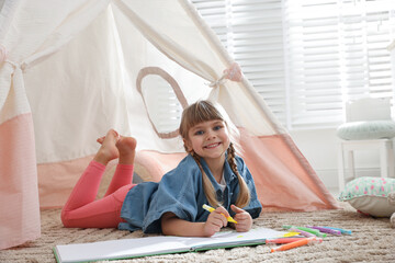 Little girl drawing in sketchbook near toy wigwam at home © New Africa