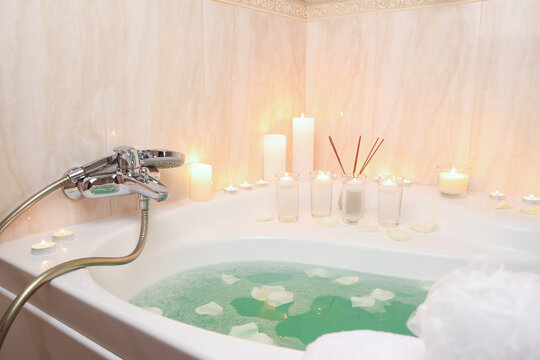Romantic atmosphere. Bathtub with burning candles, petals, air reed freshener and mesh sponge in bathroom, closeup