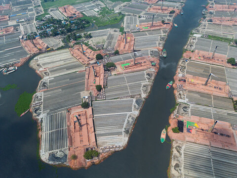 Aerial view of traditional brick kilns with tall chimneys and rows of drying bricks along the riverbank in Narayanganj, Dhaka Division, Bangladesh.