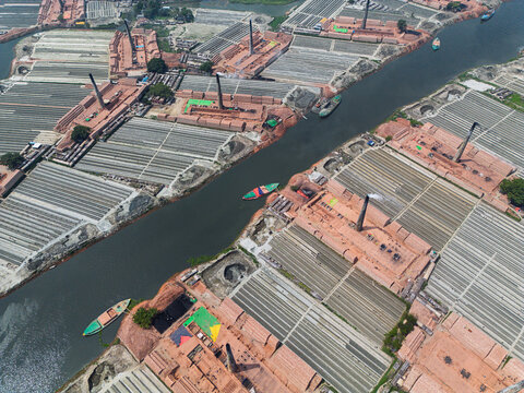 Aerial view of brick kilns with smoking chimneys and rows of drying bricks along a river with small boats Narayanganj, Dhaka Division, Bangladesh.