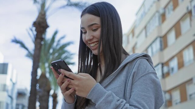 Woman smiling at smartphone and typing on a city street by palm trees and apartment balconies; youthful joy.