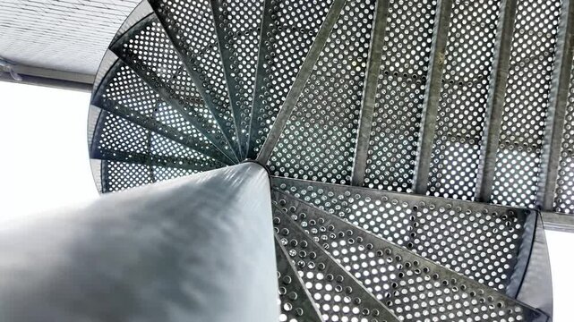 Low-angle rotating shot looking up at the center of a perforated metal spiral staircase.