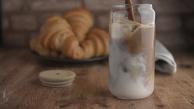 cold brew coffee or lattte, iced coffee, frappe served in a glass with pastries or croisssants on a wooden table with a rustic brick wall in the background. 