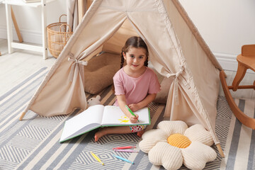 Little girl drawing in sketchbook near toy wigwam at home © New Africa