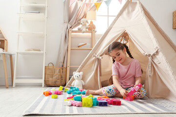Little girl playing with building blocks near toy wigwam in playroom, space for text © New Africa