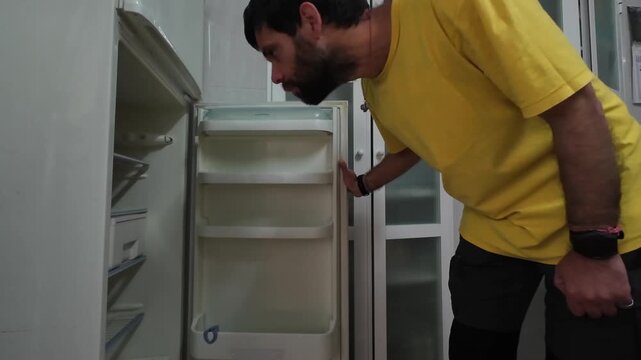 Desperate man in the kitchen opening the refrigerator door and finding no food. Concept of poverty, hunger, diet, and economic crisis
