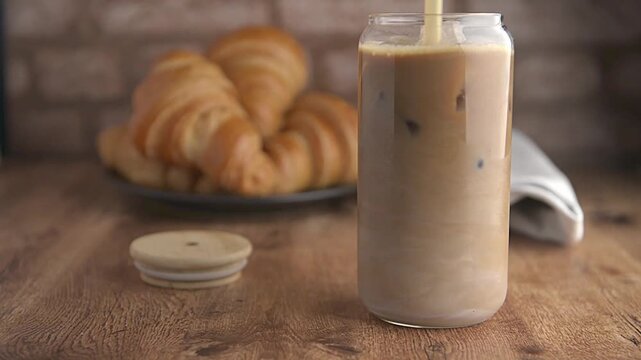 cold brew coffee or lattte, iced coffee, frappe served in a glass with pastries or croisssants on a wooden table with a rustic brick wall in the background. 