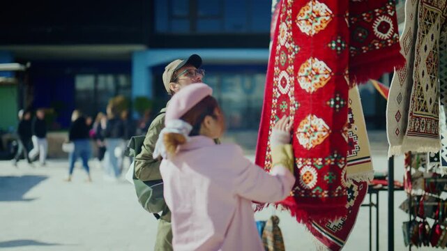 Diverse tourists are visiting a city market, browsing vibrant traditional handcrafted rugs. A couple enjoys observing colorful carpets and textiles at an outdoor stall.
