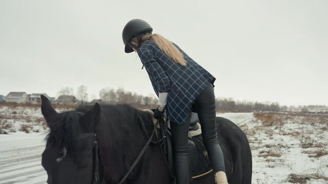 Woman mounting black horse snowy track focused training and careful saddle adjustment, rider checks stirrup and posture for lesson across flat rural road, disciplined calm and practical routine