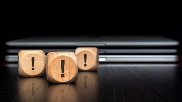 Multiple exclamation marks on wooden blocks. Blurred laptops and dark background. Cybersecurity and IT risk concept.