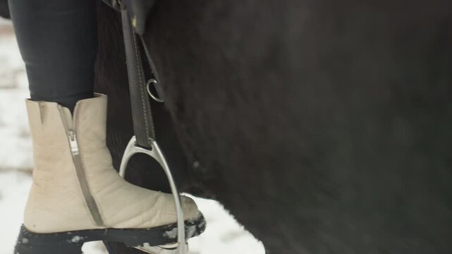 Closeup beige boot in stirrup, rider leg against black horse flank snowdusted sole and zipper, visible logo on shaft, leather saddle and stirrup leather, textured tread pressing into winter ground,