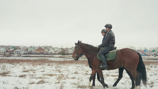 Man on horse in snowy field, lone rider undertakes slow training ride near village skyline, overcast sky, rider wearing helmet and dark jacket, green saddle, visible hoof prints in snow, calm steady