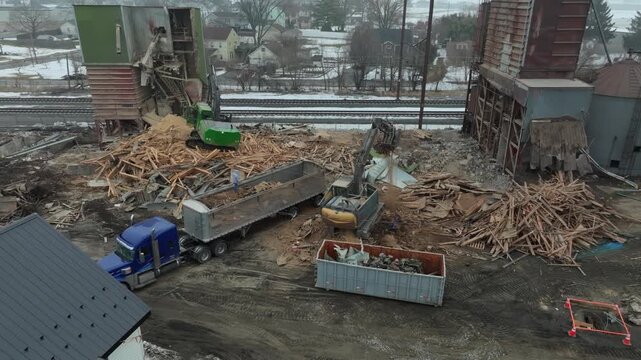 Trucks arrive at an industrial site to gather and remove large amounts of debris from a building demolition. Workers are busy clearing the area while heavy machinery operates in the background.