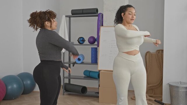 Woman stretching arms and adjusting top to expose stomach in a studio during a shared warmup session with companion; friendship calm wellness support.