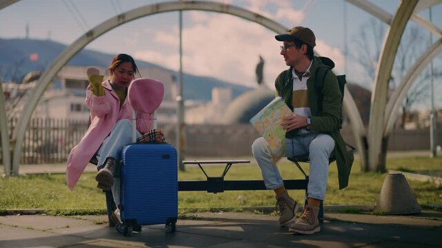 Two diverse tourists sit on a bench, consulting a map to navigate their city travels. A young couple plans their route during a relaxing urban vacation.