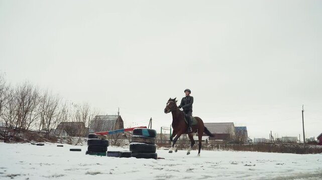 Rider on horse in snowy field, slow approach to rustic obstacle made from tires and pole, overcast sky and distant farmhouse, focused training mood with controlled pace and steady posture