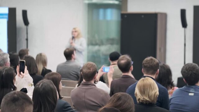 Audience listening to presenter at tech conference, people taking photos, focused crowd attending business presentation.