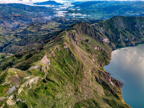Aerial view of Quilotoa Lake with steep green ridges and the volcanic crater rim overlooking the Andes mountains under a cloudy sky in Cotopaxi, Ecuador.