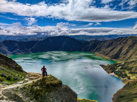 Aerial view of Quilotoa Lake with a person standing on a rocky cliff overlooking the turquoise volcanic crater lake under a dramatic cloudy sky Cotopaxi, Ecuador.