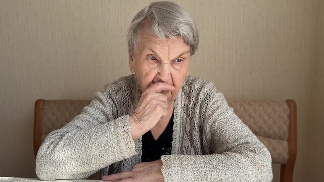 An elderly white grandmother covers her nose thoughtfully. Indoor portrait in close-up: Knitted cardigan, Gray Hair, Wrinkled facial skin. Close-up.