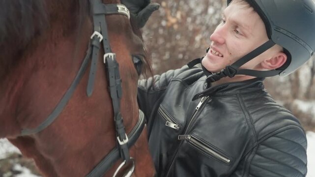 Tandem bonding grooming man with horse brushing and checking bridle, helmeted handler nuzzles chestnut mare, gentle stroking and quiet talk, winter paddock scene with snow and bare trees, intimate