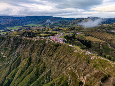 Aerial view of a small village perched on a mountain ridge overlooking deep green valleys under a cloudy sky in Cotopaxi, Ecuador.