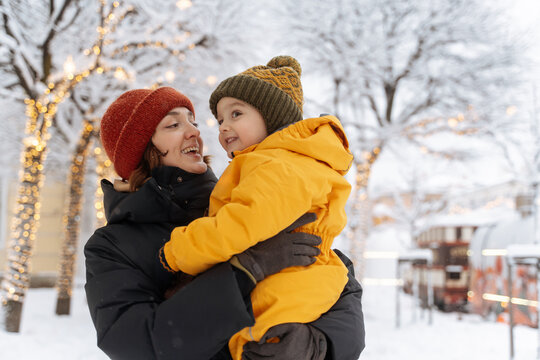 Smiling mother holding her child in a snowy park with holiday lights.