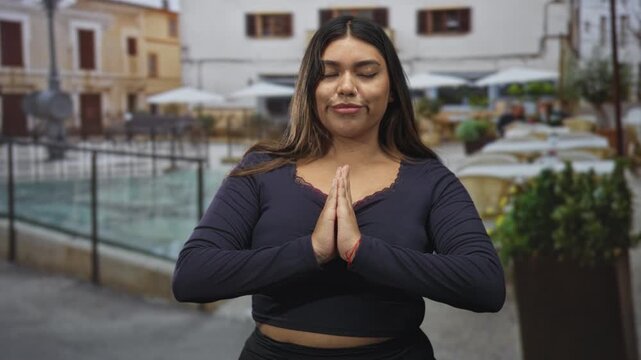 Woman pressing hands together in prayer gesture with palms visible on a street near a cafe and fountain; mindfulness serenity.