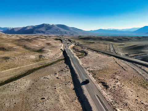 Aerial view of a long asphalt road stretching through the arid mountain landscape with a lone vehicle under a clear blue sky in Rabat Sasok-Kul, Gorno-Badakhshan, Tajikistan.