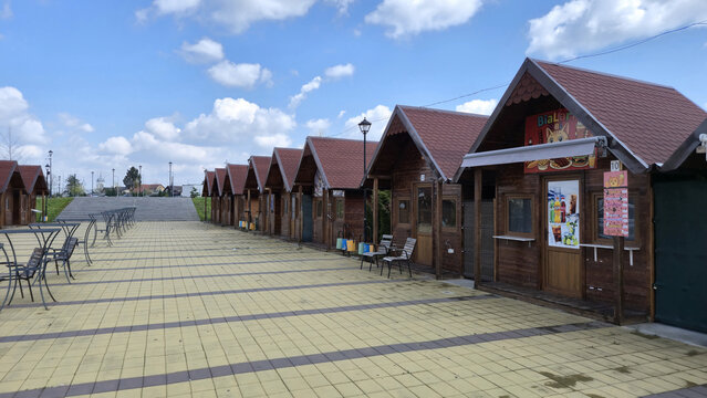 A row of wooden food stalls with red gabled roofs lines a paved pedestrian plaza under a blue sky with scattered clouds. Picture taken in April 12, 2026 in Brazii de Jos, Romania.