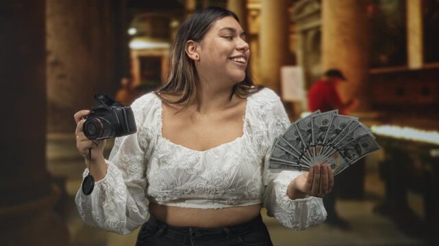 Woman holding a dslr camera and fanning us dollar bills in a grand building, smiling with exposed midriff; business confidence.