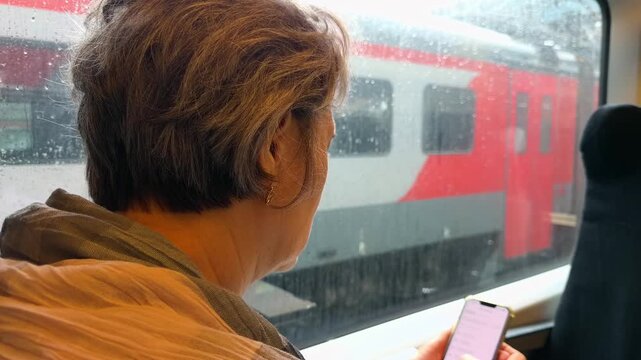 Unrecognizable Mature Woman Using Smartphone While Sitting by Rainy Window inside Passenger Train Looking at Another Railcar at Railway Station Over the Shoulder Handheld Authentic Lifestyle Shot