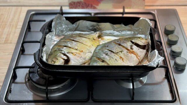 Two baked pompano on black platter overhead view, foil lined tray on gas stove burner, golden scored skin with light browning.