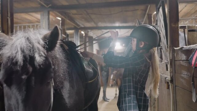 White woman saddling black horse. Helmeted rider adjusts girth and stirrup in busy barn aisle, pragmatic tandem education moment with instructor cues, leather saddle and bridle being secured, focused