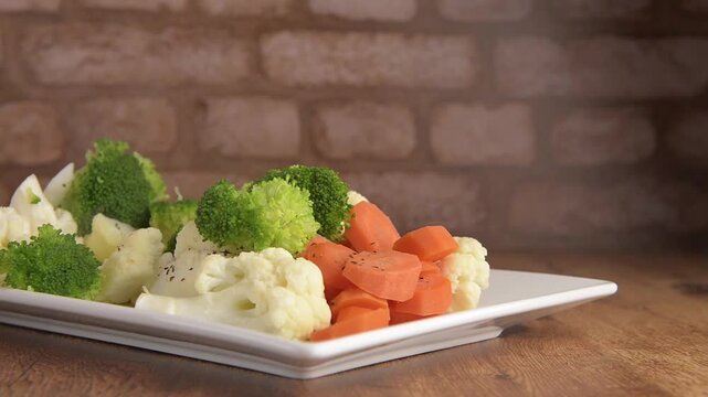 Steamed vegetables, carrots brocoli and cauliflower on a rectangular white plate to accompany meat, chicken or fish, on a rustic wooden table, with a brick wall in the background, ready to be seasoned