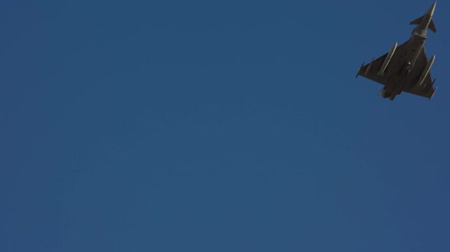 Delta wing military combat jet performing sharp rolling maneuver against clear blue sky filmed in slow motion, underside view revealing weapon station