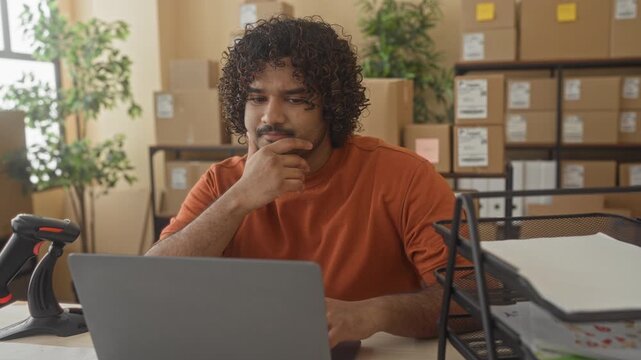 Man in orange shirt with laptop and cardboard box, hand on cheek while sorting paperwork in building; tired small business.