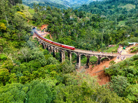 Aerial view of the Nine Arch Bridge with a red train crossing over it, surrounded by dense tropical forest and lush greenery in Demodara, Uva Province, Sri Lanka.
