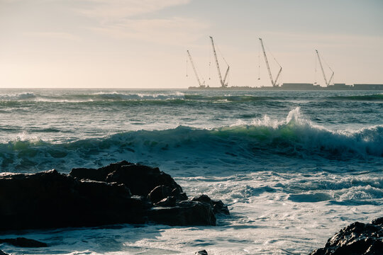 Waves Crashing Against Rocks With Cranes in the Distance