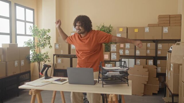 Man with laptop and cardboard box, arms raised while dancing amid stacked parcels in a building office; small business joy.