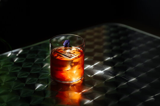 Dark cocktail with ice and flower garnish on reflective table