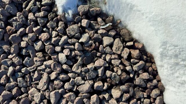 Closeup gravel meeting snow edge, detailed winter roadside texture showing small brown pebbles dusted with sparkling frost.