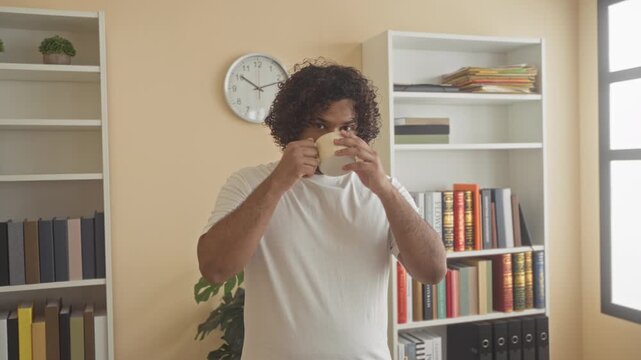 Man holding a coffee cup and sipping with both hands in a building office near bookshelves and a wall clock; contentment.