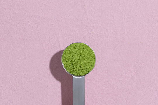 Hand Holding Measuring Spoon with Green Moringa Powder on pink