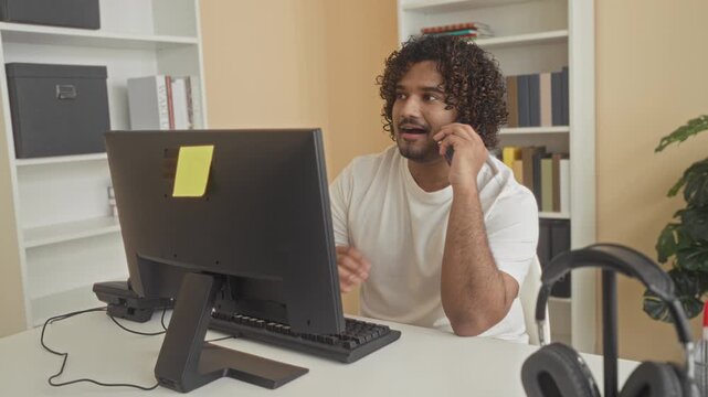 Man holding smartphone to ear and typing at computer monitor at office desk with sticky note on screen; productive remote work.