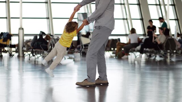 Father twirls joyful toddler around, creating airborne giggles and carefree fun. Playful swirl in waiting area of large airport terminal, with public lounge visible blurred in background. Slow motion