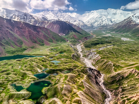 Aerial view of Tulpar Lake with turquoise water surrounded by green rolling hills and snow-capped Pamir Mountains in Alay District, Osh Region, Kyrgyzstan.