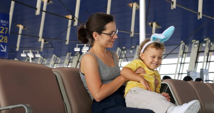 Mother and her young son, wearing bunny ears for Easter, sit in airport terminal. Baby boy bounces playfully on her lap, giggling as she tilts him to side, sharing joyful moment while waiting