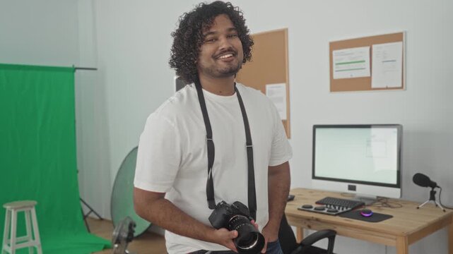 Young indian man photographer holding camera with hand, strap around neck, smiling and hand on hip in studio; confidence creativity.