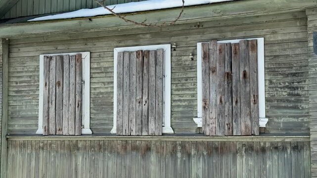Boarded winter farmhouse facade with snow, weathered wooden shutters and peeling paint, cracked attic window in gable, pale blue.